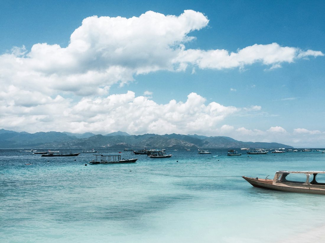 Small wooden boats anchored in the clear turquoise water off the Gili islands, Lombok's coastline in the distance.
