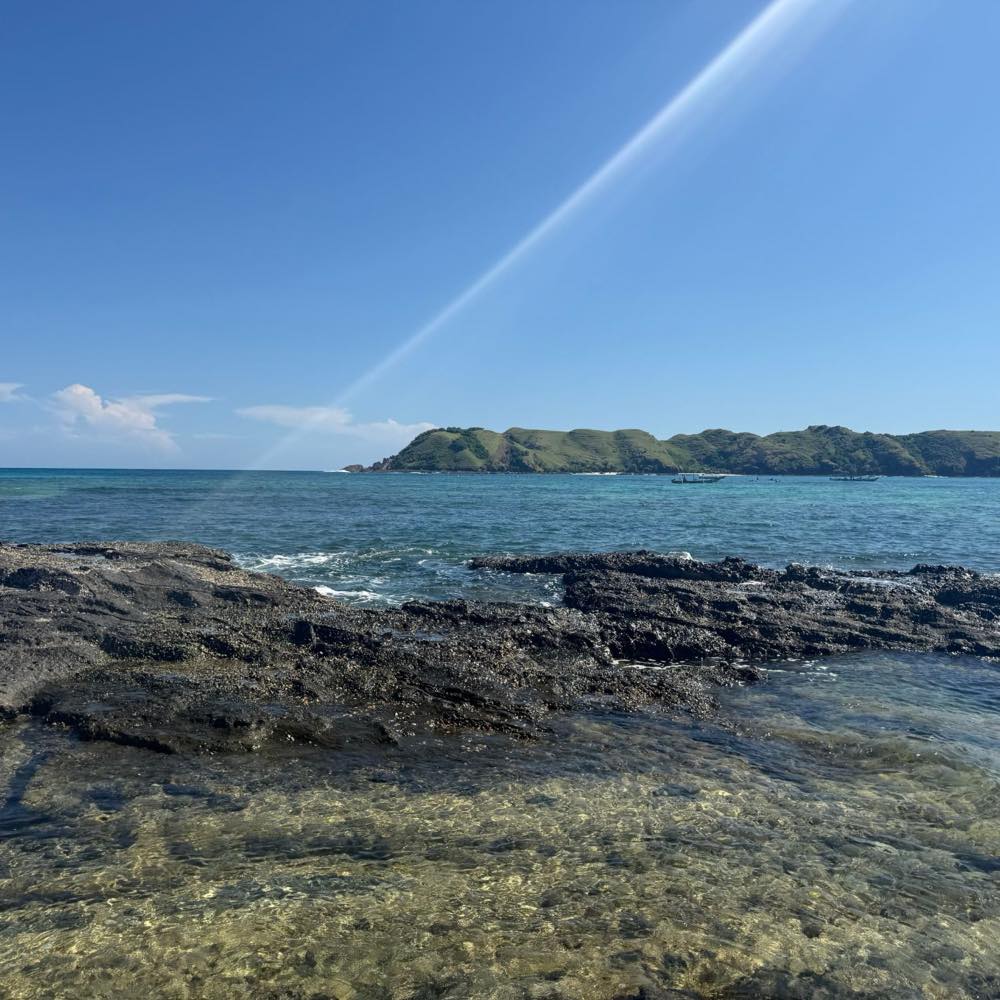 Turquoise water over a rocky shore on Lombok, with boats moored off a green headland in the distance.